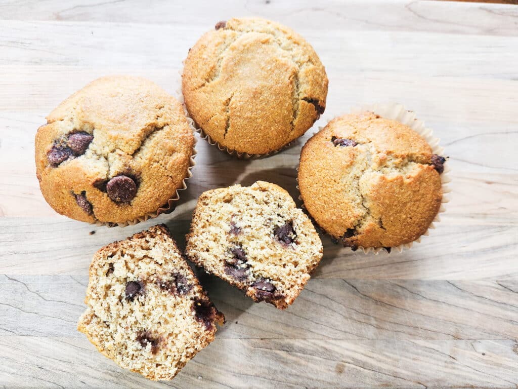 fresh milled flour sourdough chocolate chip muffins presented on a cutting board with 3 whole, puffy muffins, and 1 muffin cut in half, displaying squishy dough