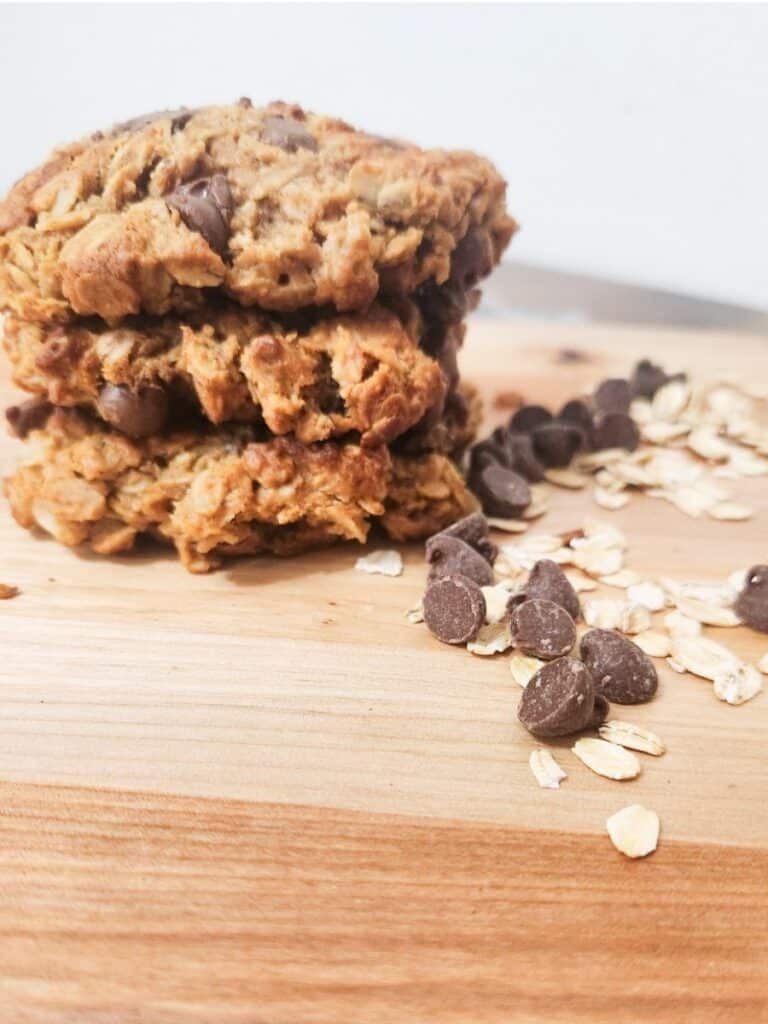 3 giant monster cookies displayed on a cutting board, stacked on top of each other with oats and chocolate chips surrounding the stack. cookies look soft and chewy